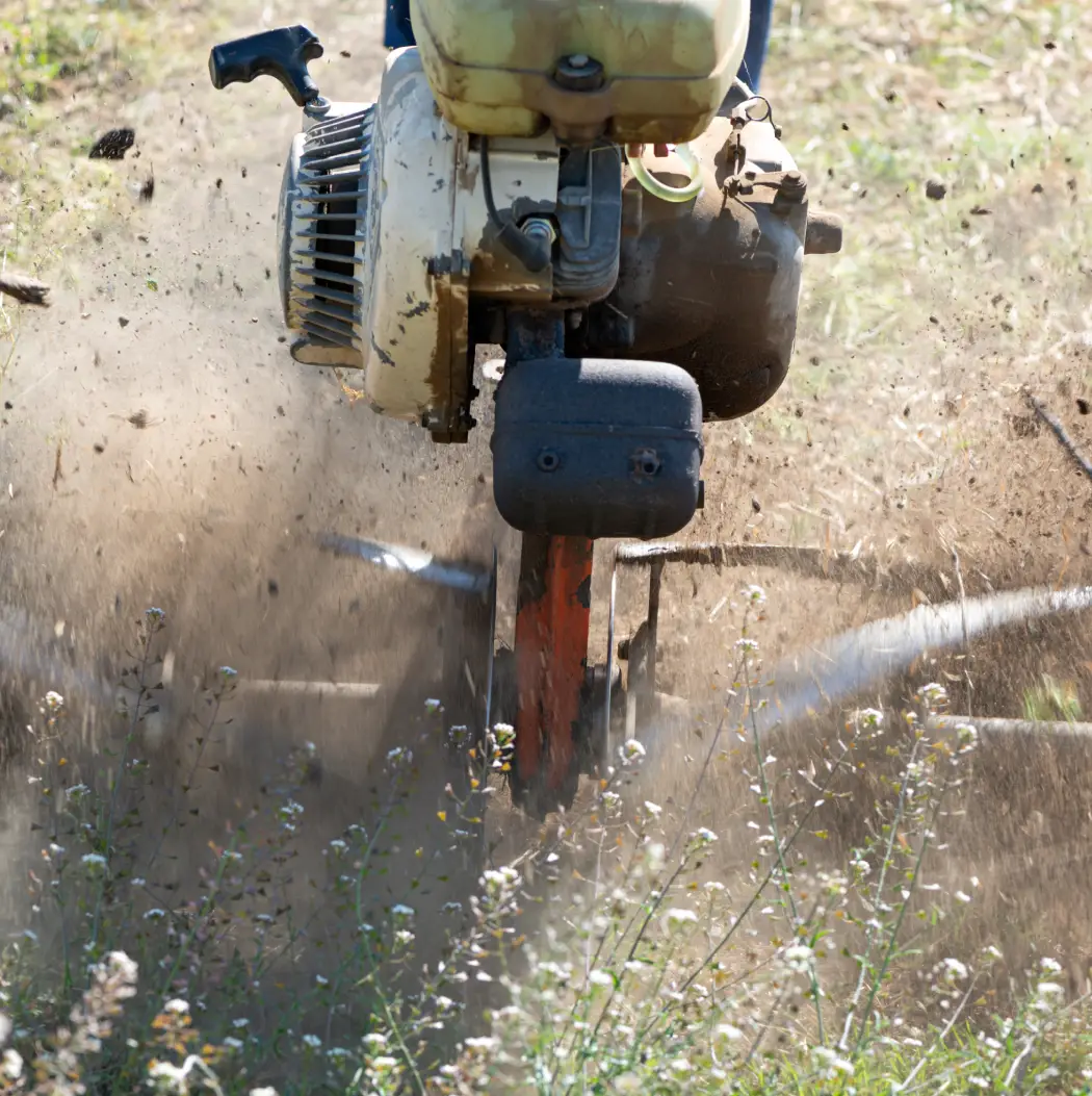 A powerful tiller digs into the earth, creating a cloud of dust while aerating the soil. Surrounding greenery enhances the rugged nature of this agricultural scene, where machines meet nature in harmony.